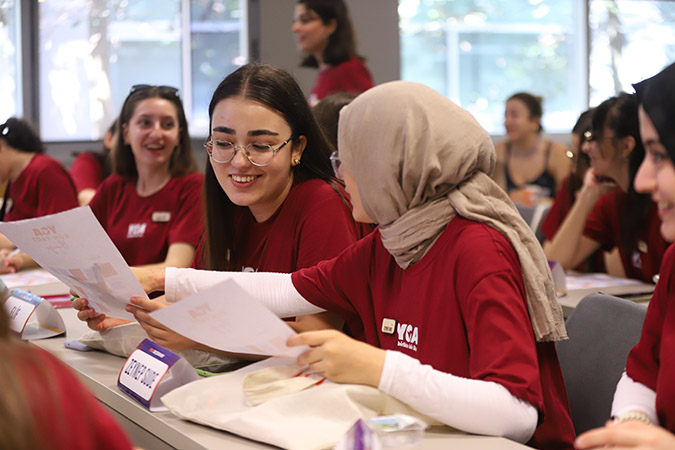 Female wearing tan hajib, crimson Young Guru Academy T-shirt over white long sleeved shirt, holding a paper in left hand while pointing to something on a paper held by a female seated to her left with uncovered long brown hair, both in a classroom surrounded by several other females wearing similar T-shirts.