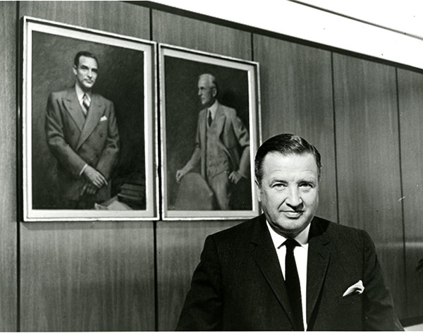 Henry Ford II in front of portraits of his father, Edsel Ford, and his grandfather Henry Ford, in a wood paneled room.