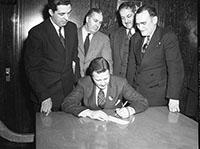 Four suited males look on as Henry Ford II signs a check for the American Red Cross. 