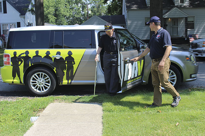 A person holds the door of an SUV vehicle while a person wearing WORLD WAR II VETERAN embroidered hat steps out with a cane