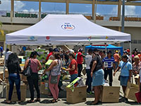 Five people in line getting Coleman gas stoves and gas canisters under a Ford CENTRO DE MOVILIDAD SOCIAL pop-up tent with numerous others and staff in the background near other tents.