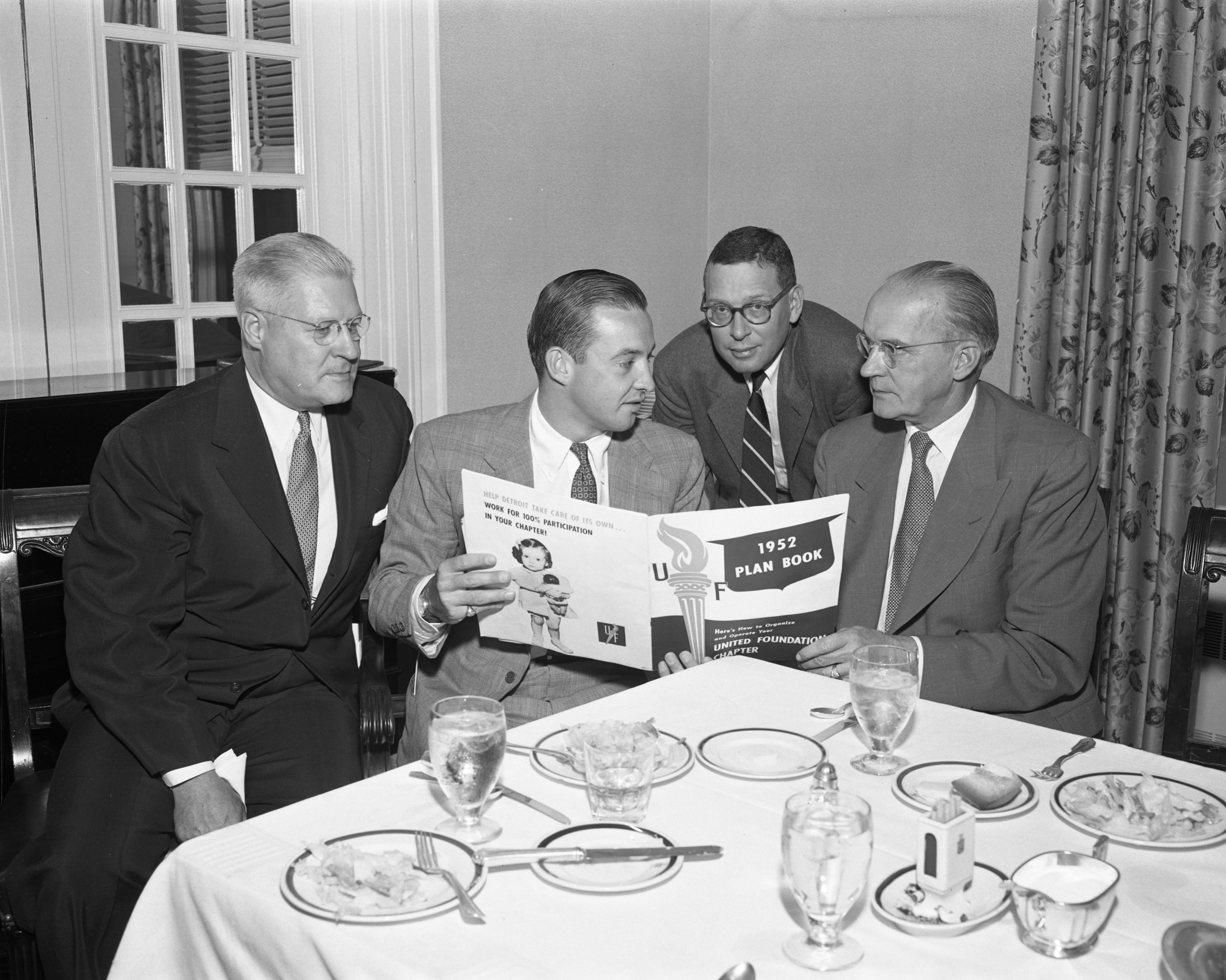  William C. Ford chairman of United Foundation Industrial Group with trio of males, all in suits and ties, seated at table after a meal with dishes still on the table, as he holds 1952 UF Plan Book.